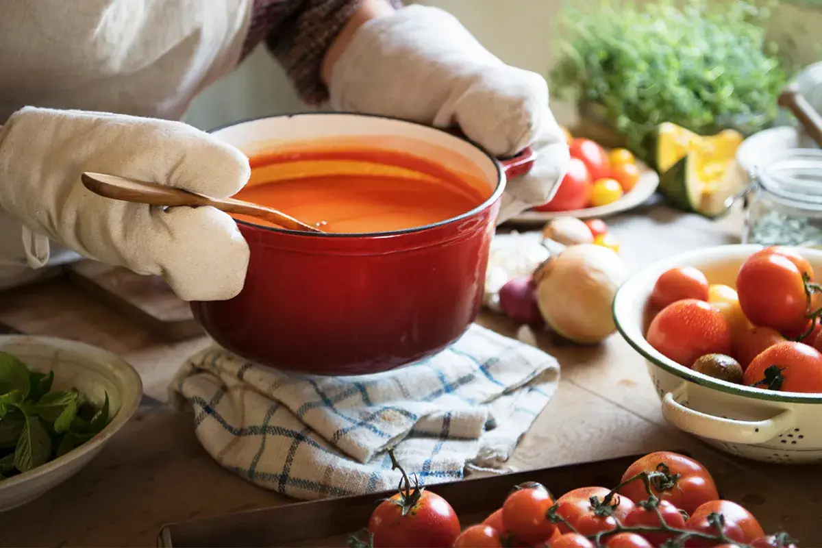 A person wearing oven mitts holds a red pot of tomato soup on a kitchen towel, surrounded by fresh tomatoes and vegetables on a wooden table.