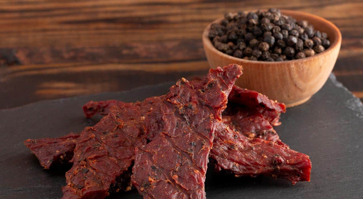 Slices of beef jerky on a slate board with a wooden bowl of black peppercorns in the background.