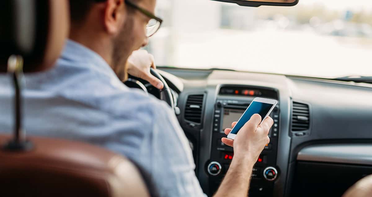 A man in a car is looking at his smartphone while sitting in the driver's seat. The dashboard and steering wheel are visible.