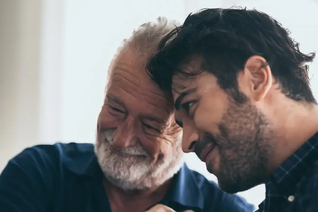 An older man and a younger man sit closely together, smiling with their heads touching, suggesting a moment of warmth and connection.
