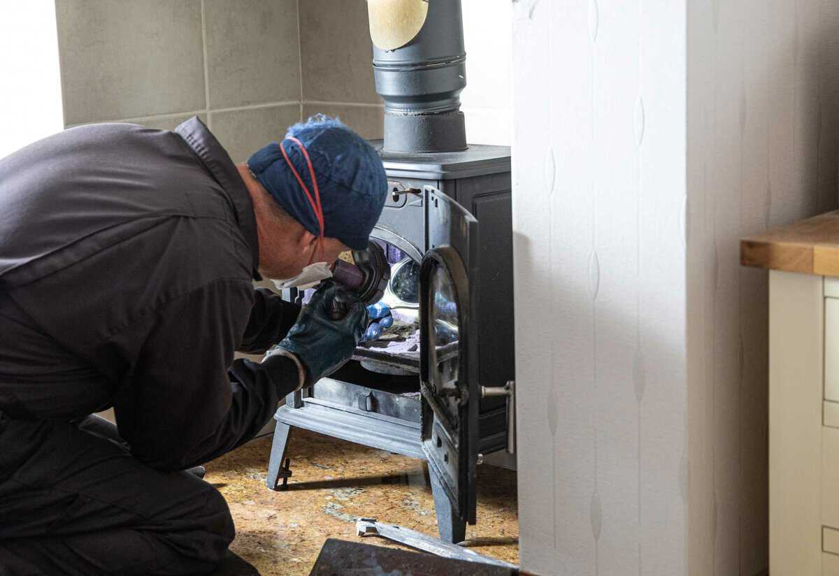 A person in protective gear inspects the inside of a wood-burning stove, holding a flashlight to illuminate the area.