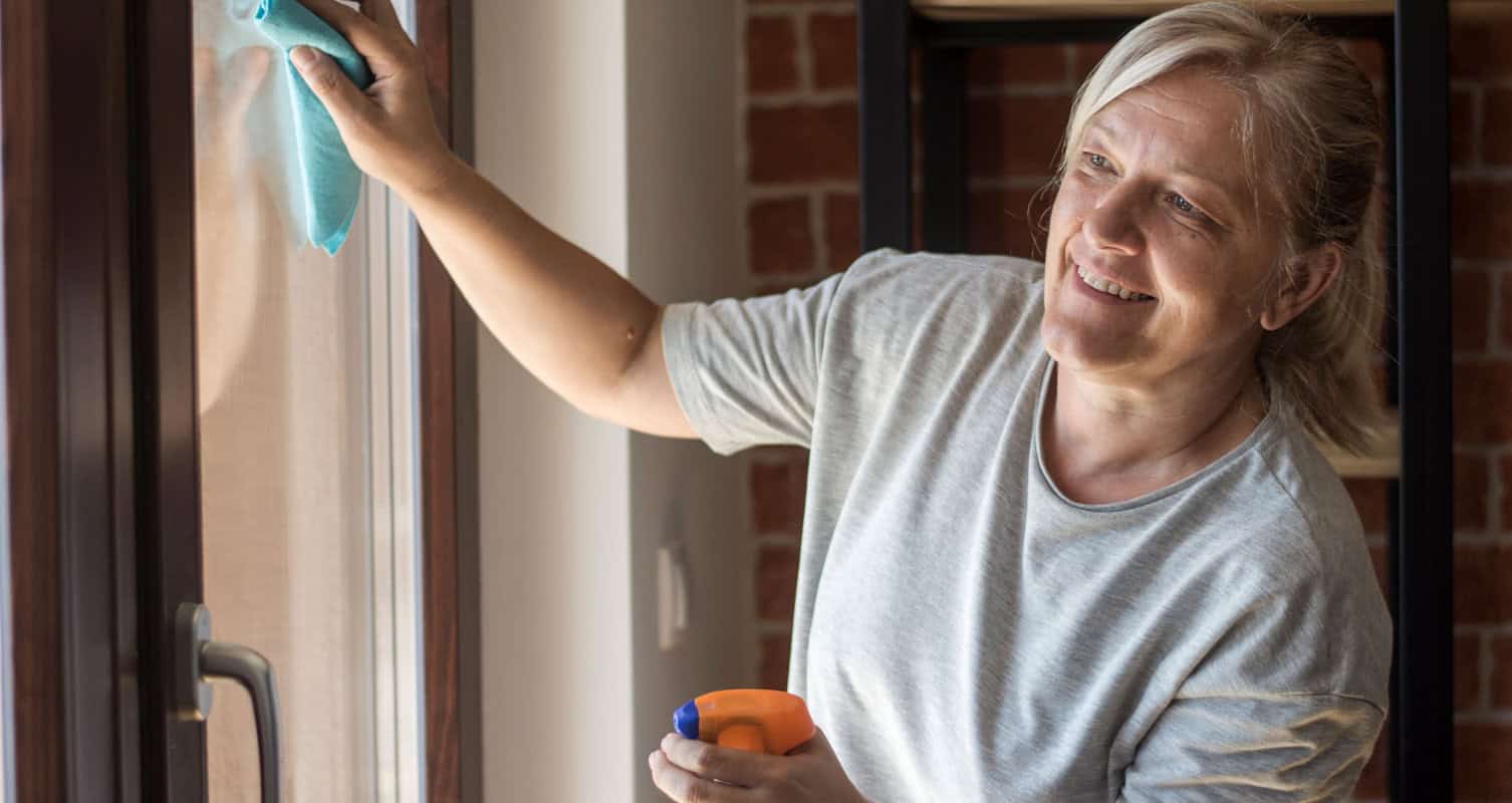 Woman cleaning a window with a cloth and spray bottle, smiling.