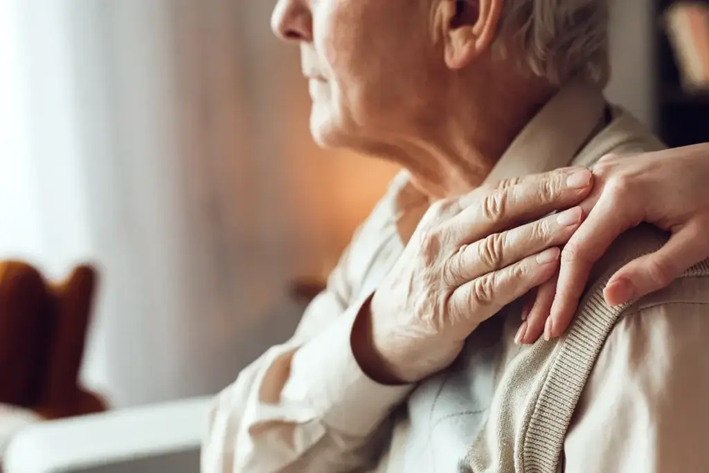 An elderly person sits indoors with a younger hand resting on their shoulder in a comforting gesture.