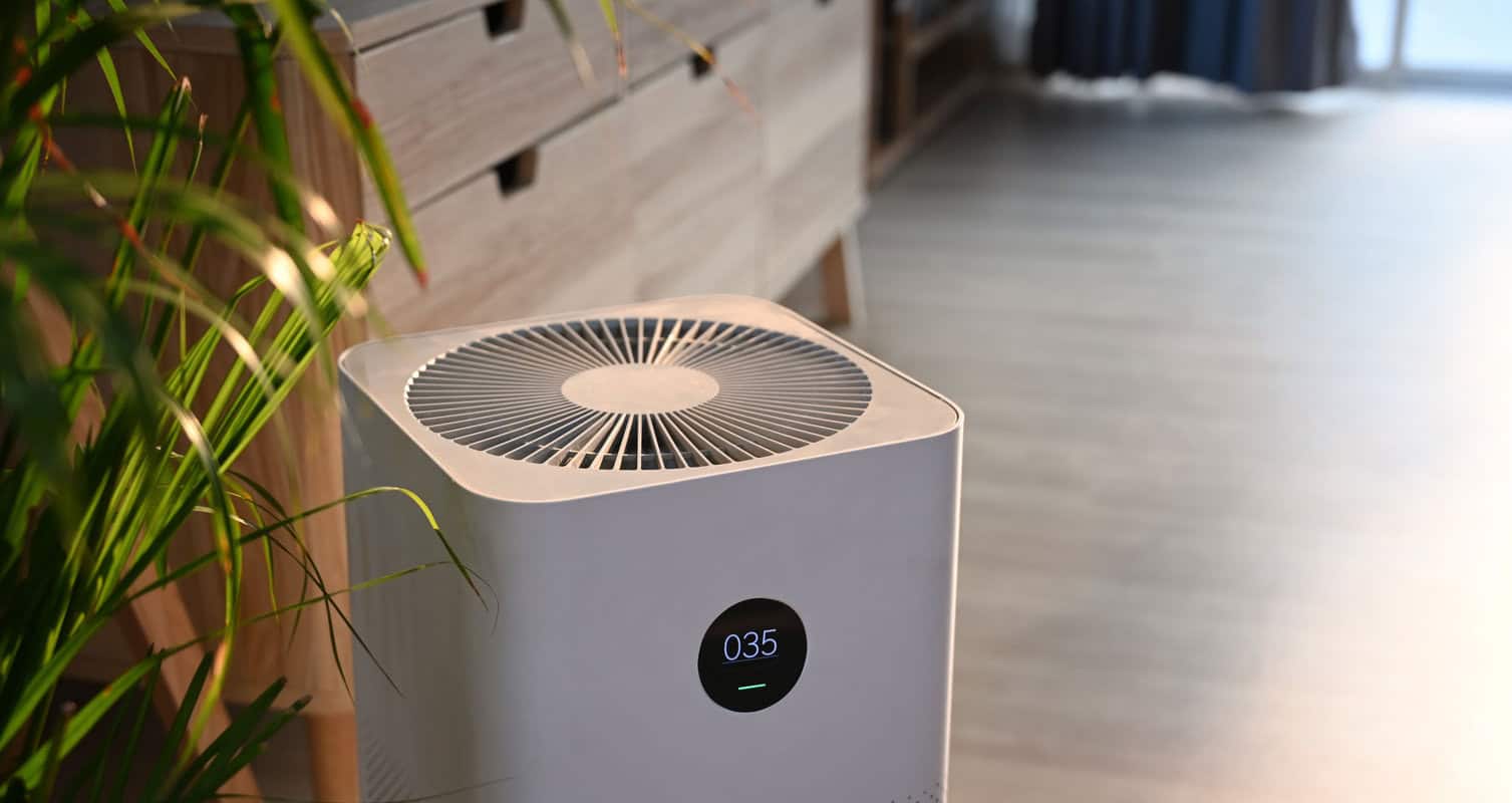 A modern air purifier with a digital display reading "035" is placed on a wooden floor near a green plant and a wooden cabinet.