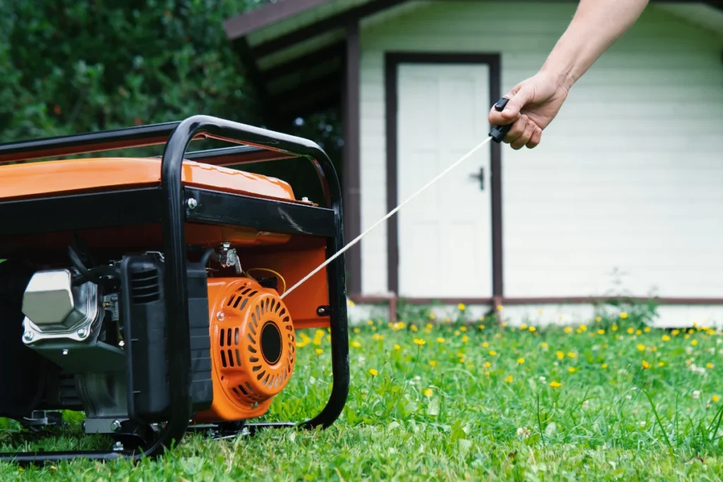 A person pulls the starter cord of an orange portable generator on grass in front of a small white building.