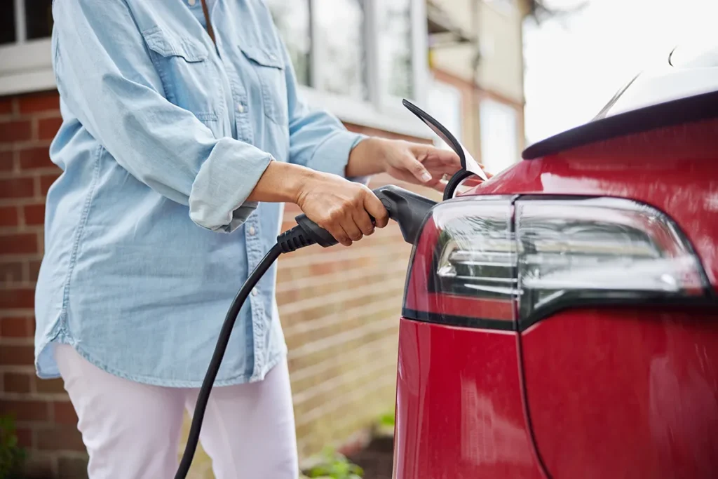 Person in a light blue shirt plugs a charging cable into a red electric car parked outside near a brick building.