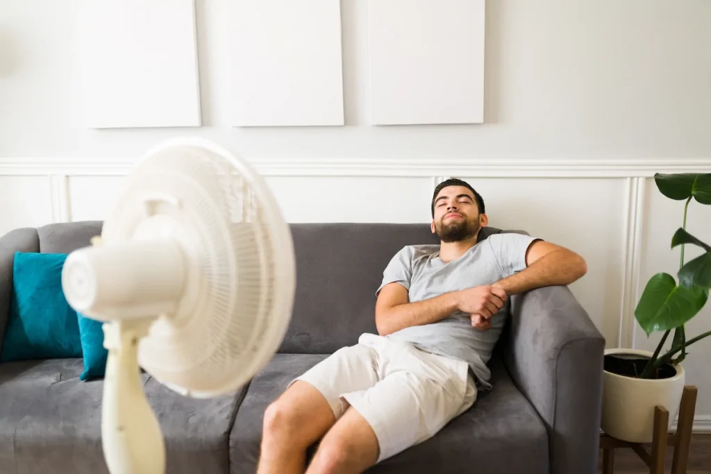 Man sitting on a gray sofa with eyes closed, facing an oscillating fan in a living room with minimal decor and a potted plant.