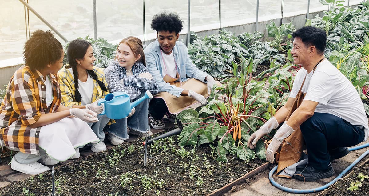 A diverse group of people gardening in a greenhouse. Some are kneeling, tending to plants with tools and watering can, while one person is holding a garden hose.