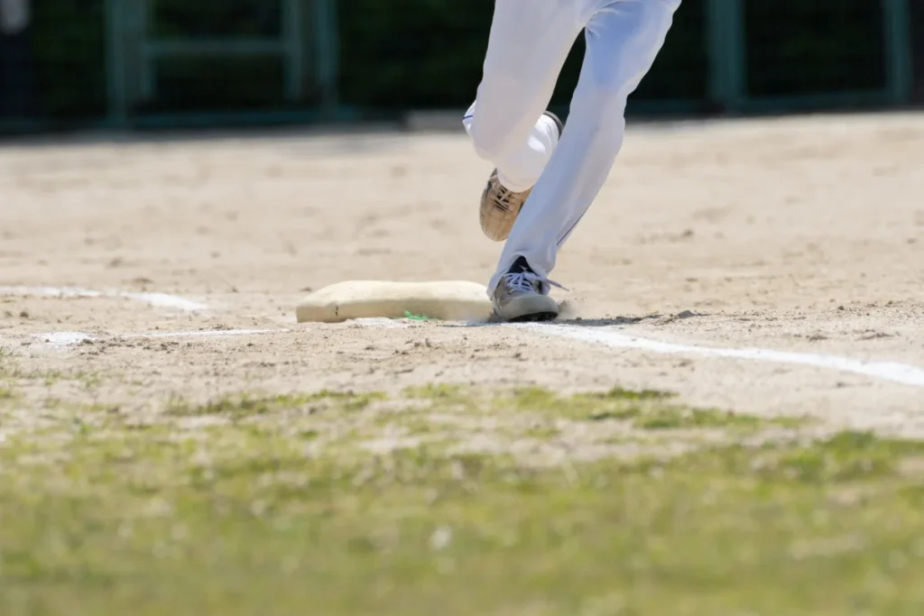 A baseball player in white pants and cleats steps on a base on a sandy field, with only the lower legs visible.