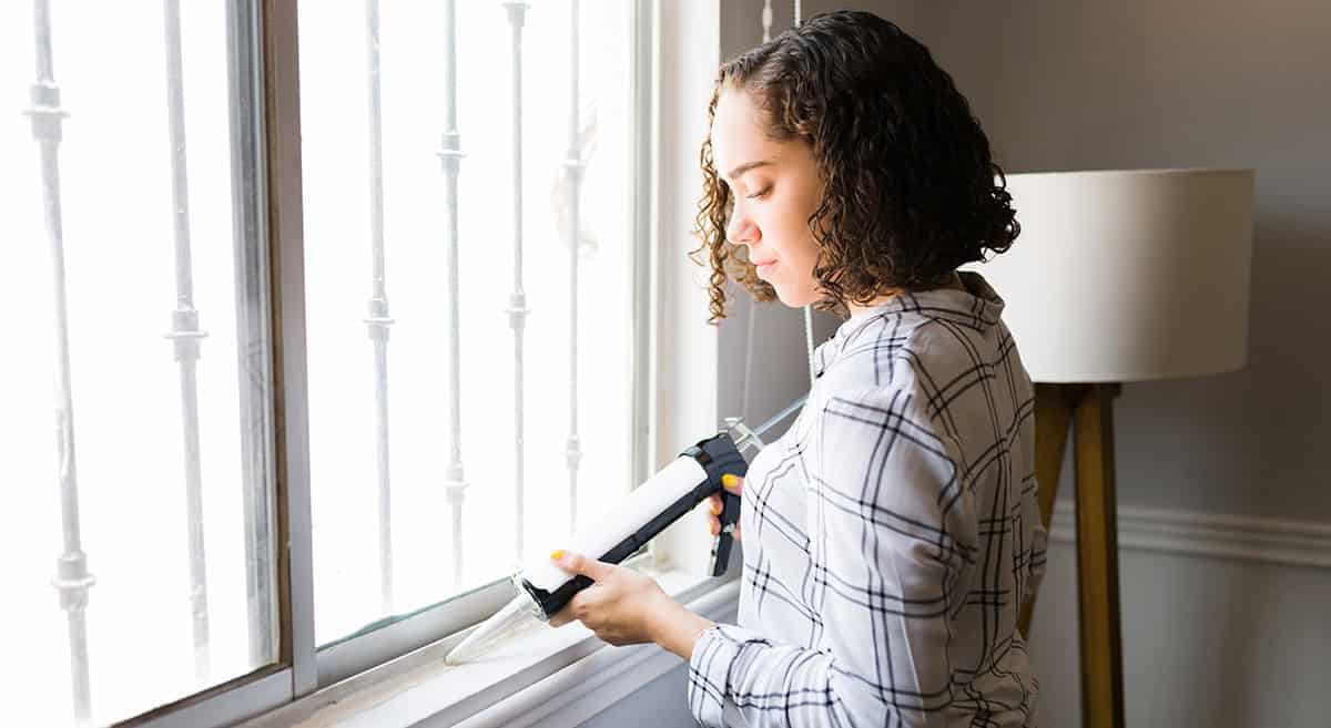 A person applies sealant to a window frame inside a room with a lamp and barred window.