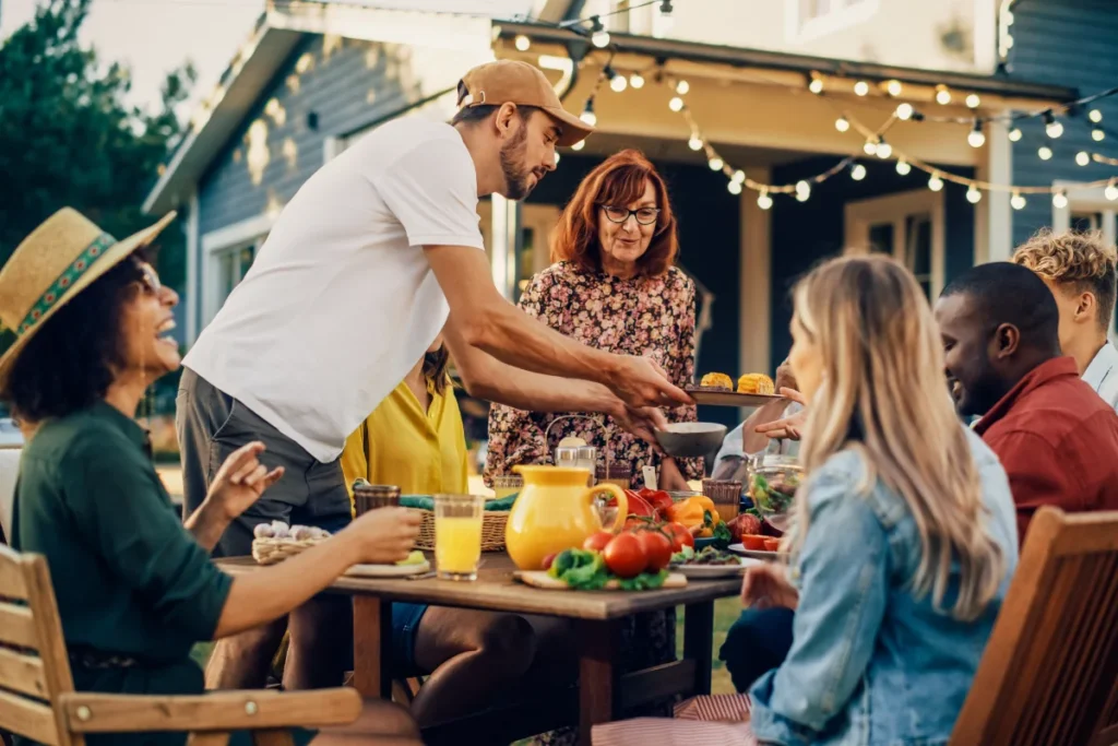 A group of people sit around an outdoor table sharing a meal, with drinks and food, in a backyard decorated with string lights.