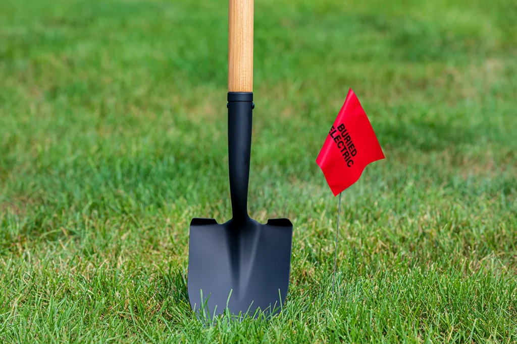 A shovel with a wooden handle is stuck in green grass next to a small red flag labeled "BURIED ELECTRIC LINE.