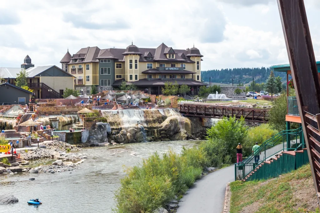 A riverside scene with a large hotel building, a pedestrian bridge, people walking on a path, and greenery along the water under a cloudy sky.