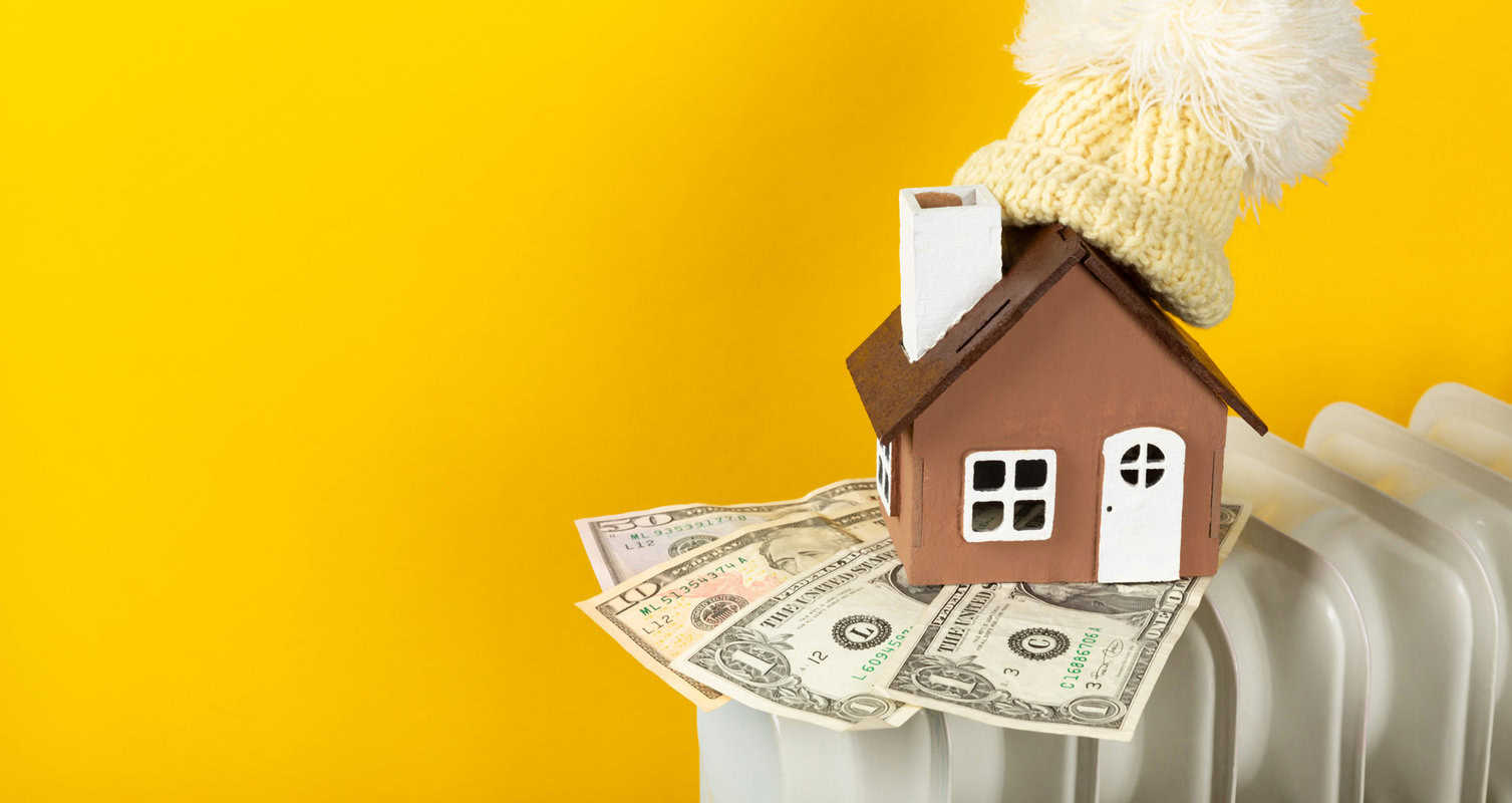 A small house model with a knitted hat on top, sits on dollar bills placed over a radiator, against a yellow background.