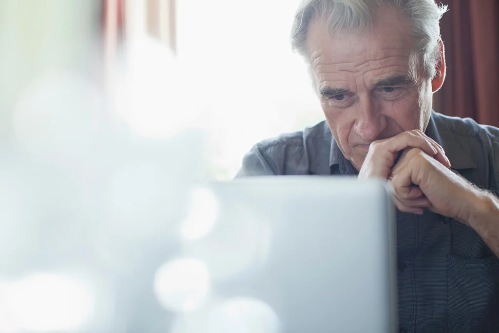 Older man with gray hair sits at a table, looking intently at a laptop screen with his hands resting under his chin.