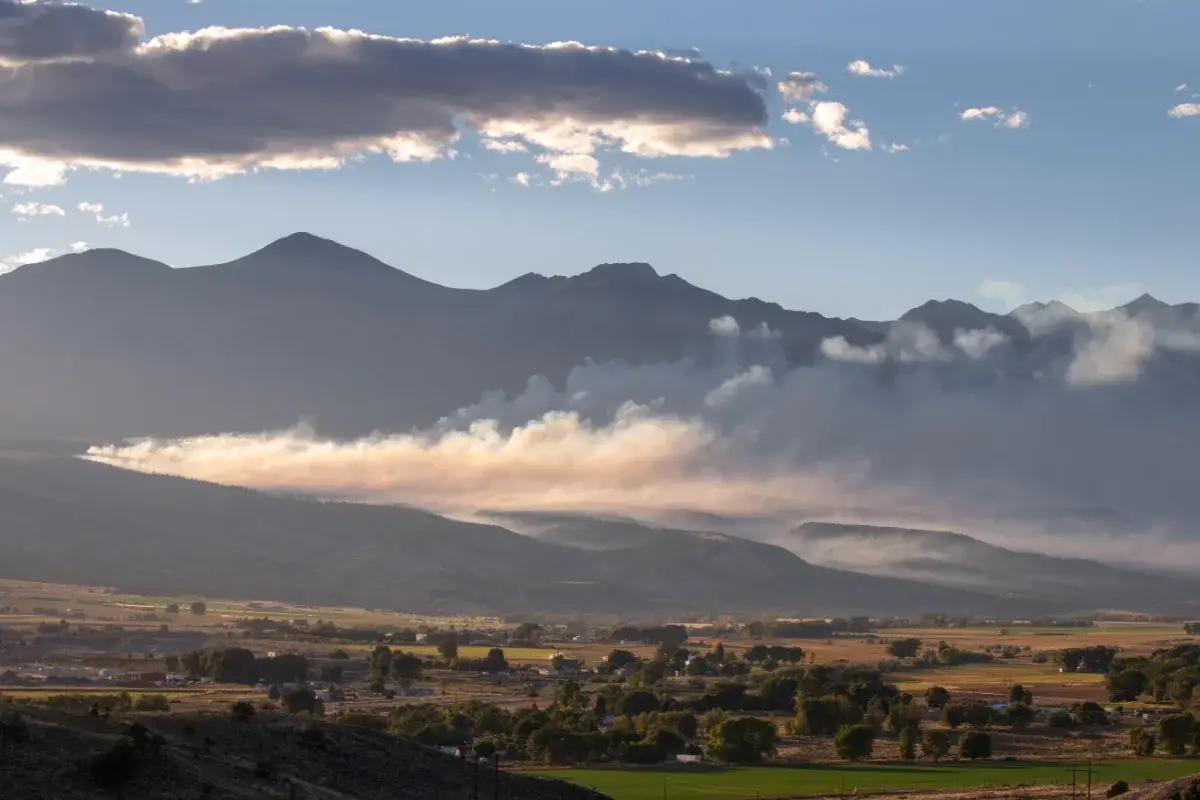 Smoke drifts across a rural valley with houses and fields, while mountains and clouds rise in the background under partly cloudy skies.