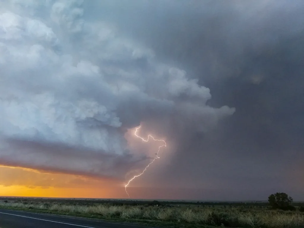 A bolt of lightning strikes from a dark, dense cloud over an open landscape at sunset, with the sky showing shades of orange and gray.