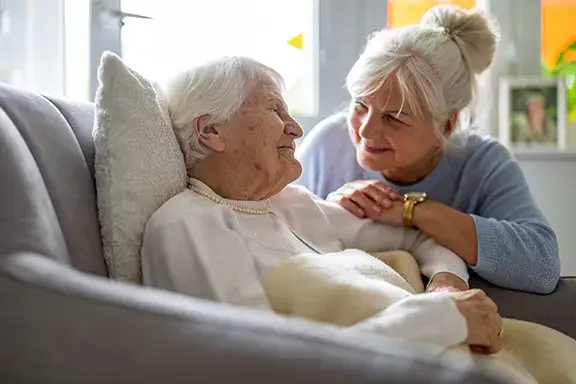 An elderly woman lies on a sofa under a blanket while a middle-aged woman sits beside her, smiling and holding her arm in a bright room.