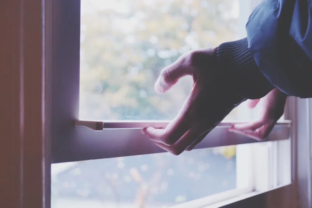 A person’s hands are shown opening a window with a white frame, letting in natural light from outside.