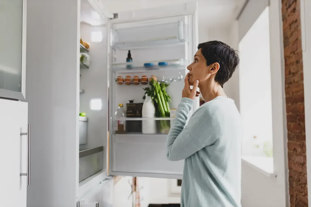 A person stands in front of an open refrigerator, looking inside thoughtfully with a hand on their chin.