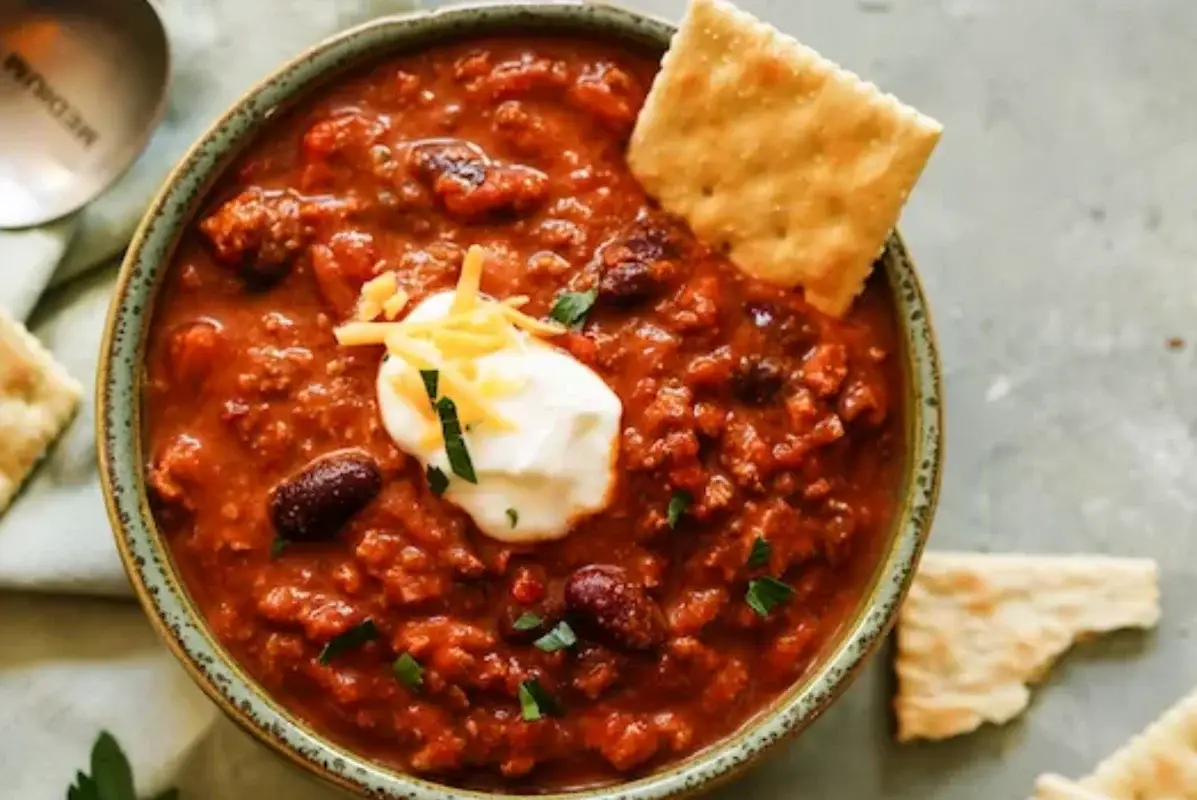 A bowl of chili topped with sour cream, shredded cheese, and parsley, accompanied by saltine crackers on the side.