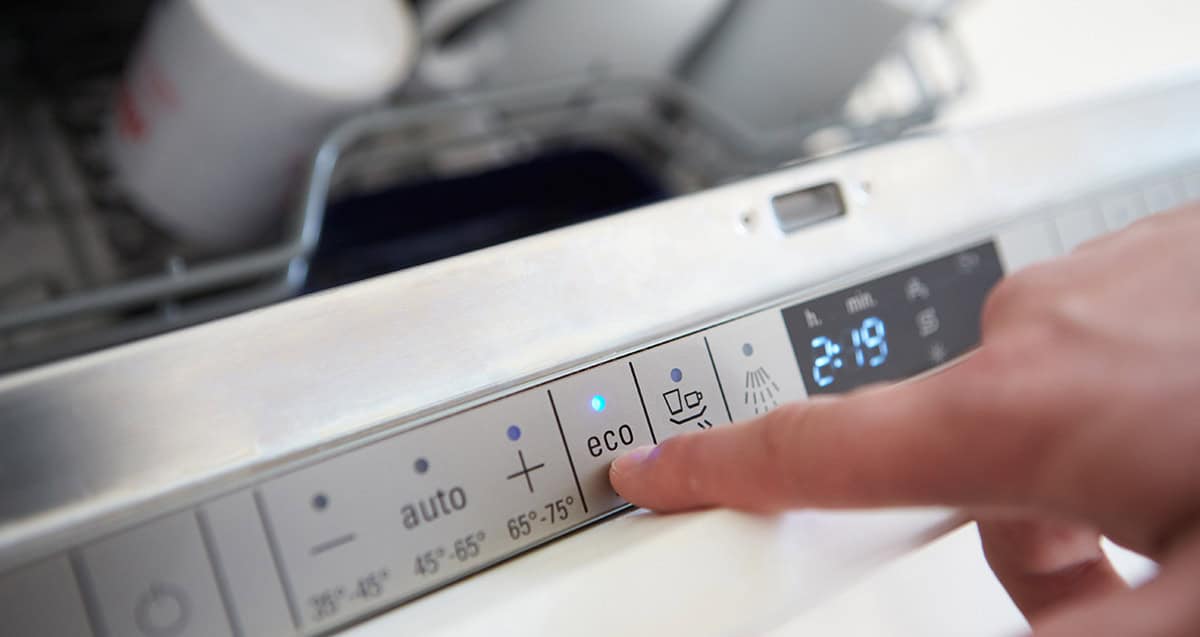 Finger pressing the eco mode button on a dishwasher control panel, with the appliance door slightly open and some dishes visible inside.