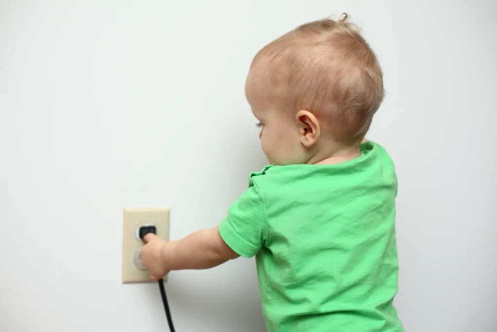 A toddler in a green shirt reaches toward an electrical outlet to touch a plugged-in cord against a plain white wall.