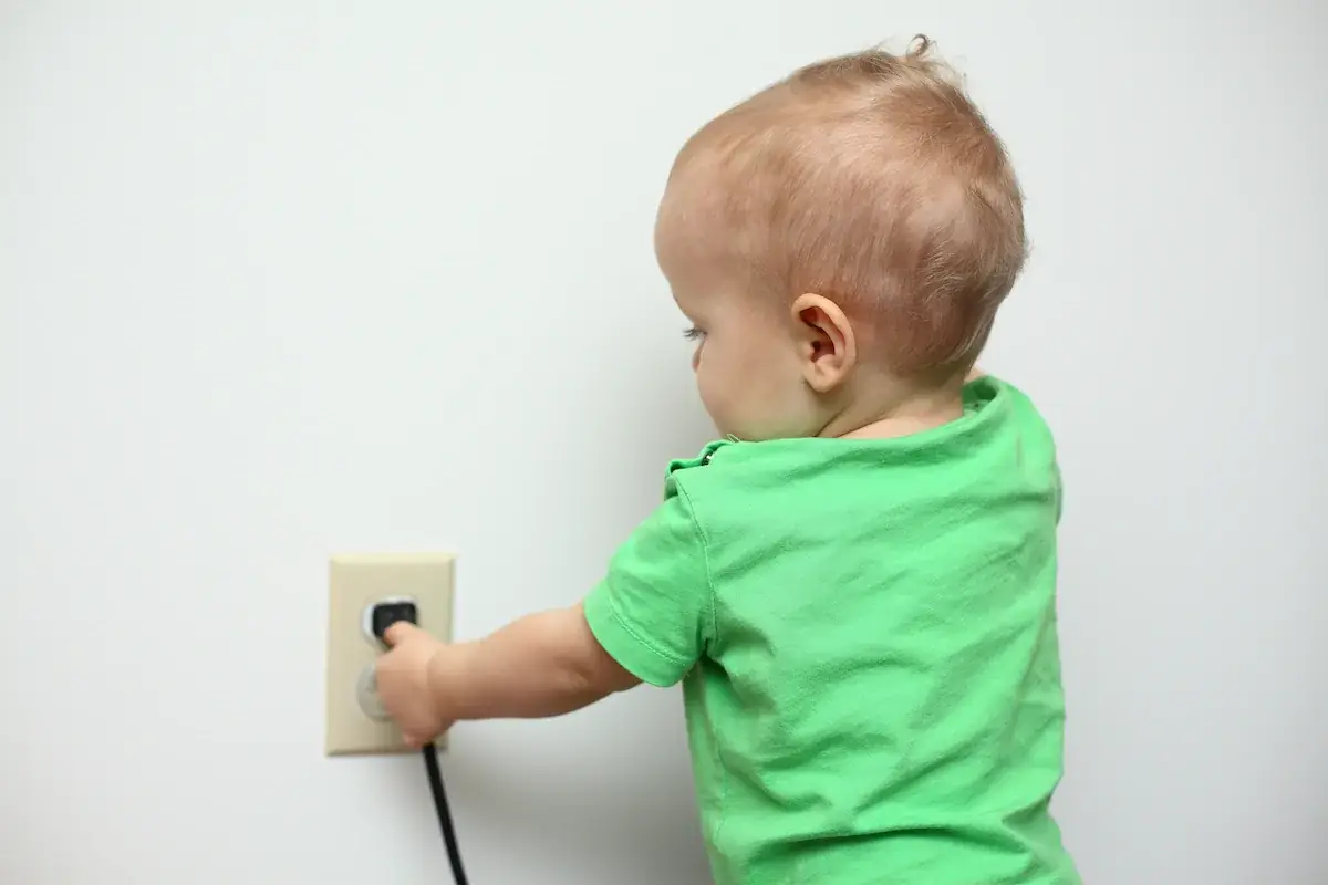 A toddler in a green shirt reaches toward an electrical outlet to touch a plugged-in cord against a plain white wall.