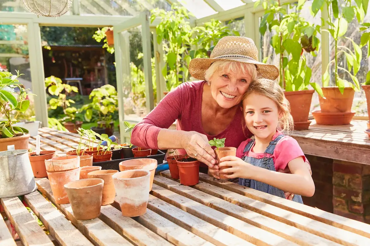 An older woman and a young girl smiling while planting seedlings together in a greenhouse filled with potted plants.