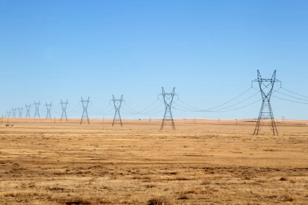 A row of high-voltage power transmission towers stretches across a flat, dry, grassy plain under a clear blue sky.