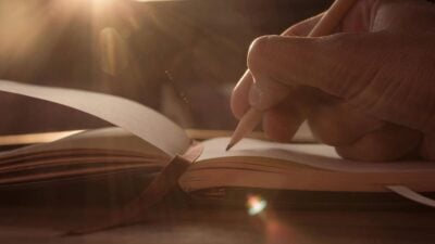 Hand writing in a notebook with a pencil, sunlight streaming from the top left.