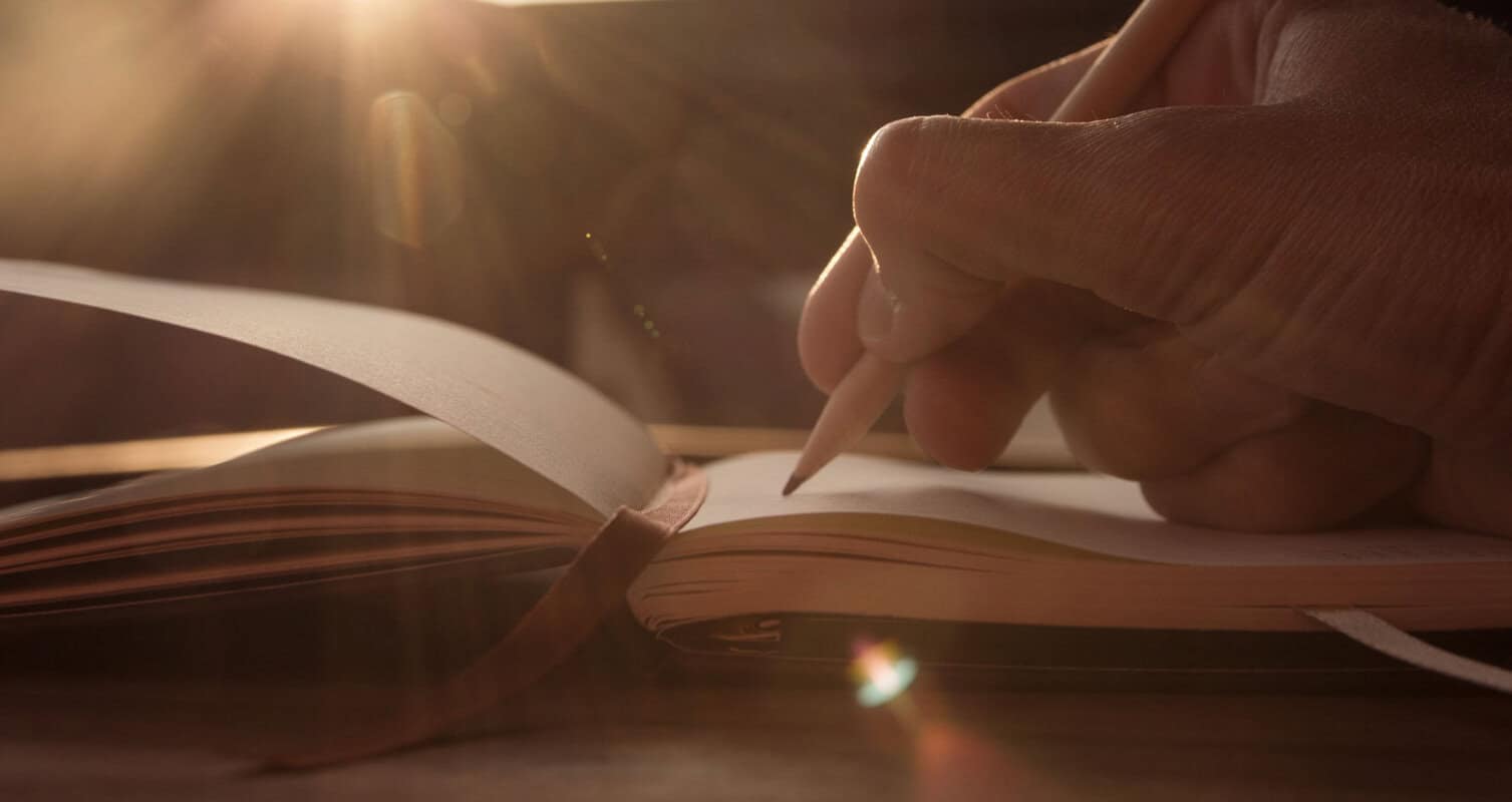Hand writing in a notebook with a pencil, sunlight streaming from the top left.