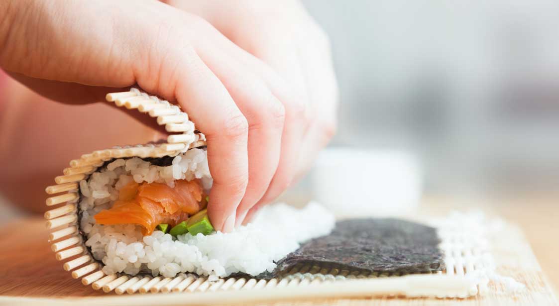 Hands rolling sushi with rice, salmon, and vegetables using a bamboo mat.