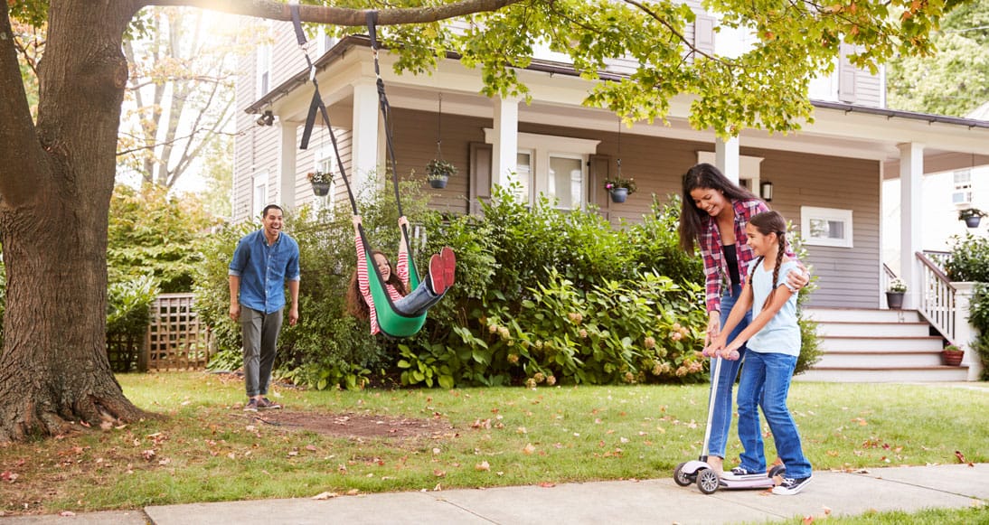 Two adults and two children in a suburban yard; one child swings, another rides a scooter with an adult's help, while another adult watches.