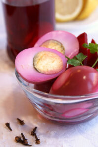 Quickly Pickled Pink A glass bowl with pink pickled eggs and parsley, surrounded by cloves, with a jar and lemon slices in the background.