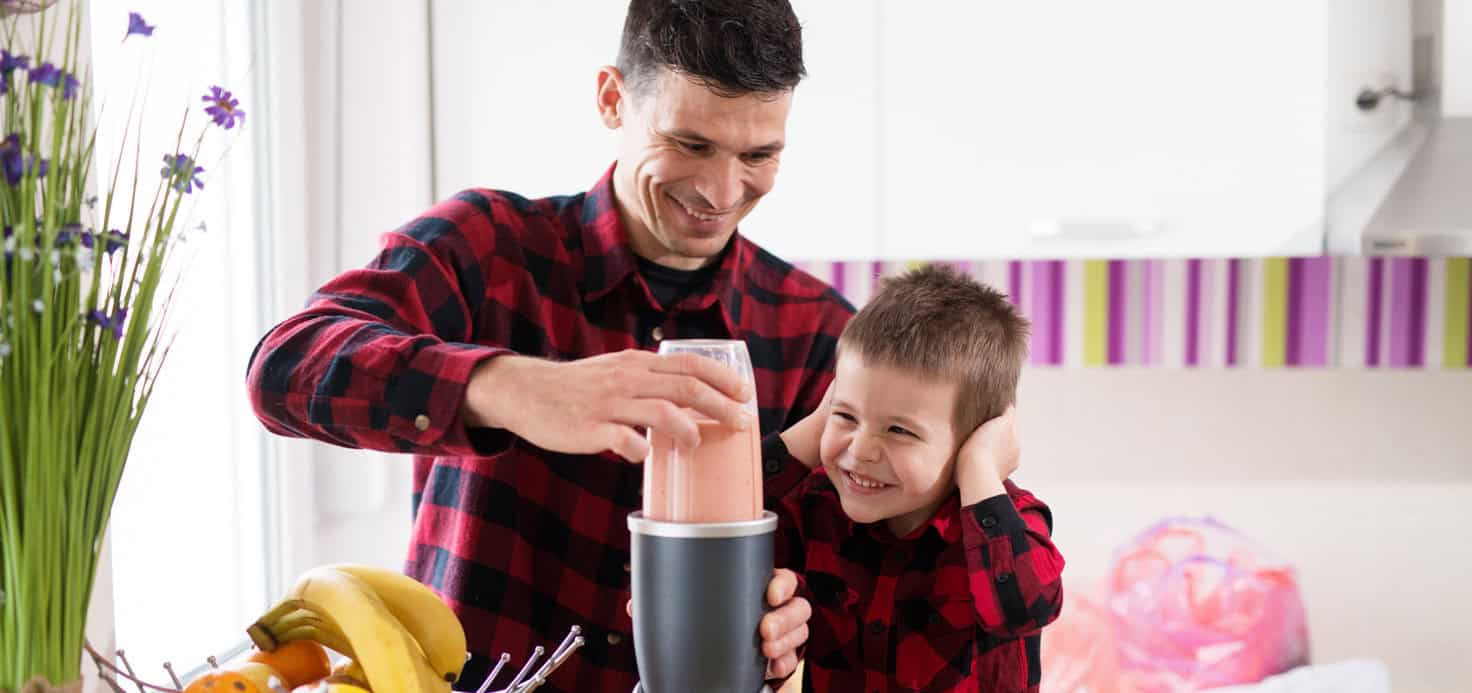 A man is using a blender while a young boy covers his ears in a kitchen. They are both wearing red plaid shirts.