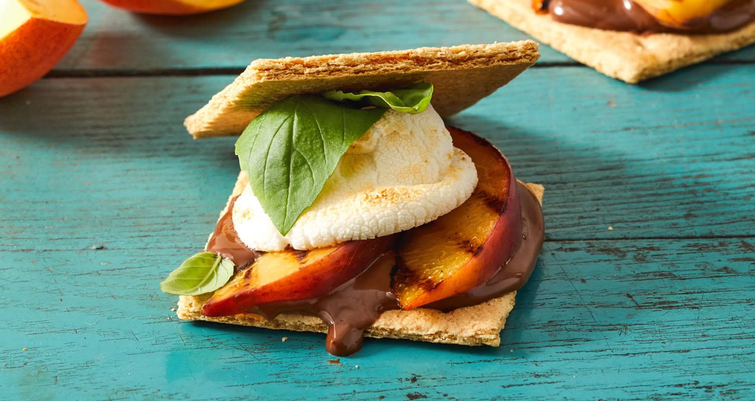 A s'more with grilled peaches, melted chocolate, a toasted marshmallow, and basil leaves between two graham crackers, on a blue wooden surface.