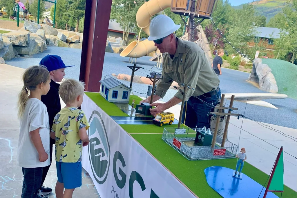 An adult in a hard hat demonstrates a model electrical grid to three children at an outdoor event with playground equipment in the background.