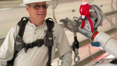 A worker in safety gear stands next to a red elf doll perched on a metal structure, with the Grand Valley Power logo in the corner.