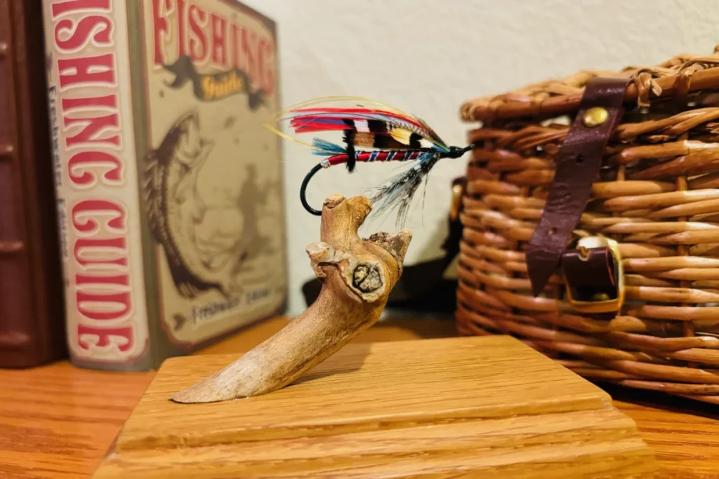 A decorative fishing fly is displayed on a wooden stand, with a fishing guidebook and a woven basket in the background on a wooden surface.
