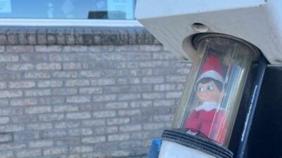 Elf doll inside a transparent tube on a drive-thru intercom, with an "Open" sign in the background.