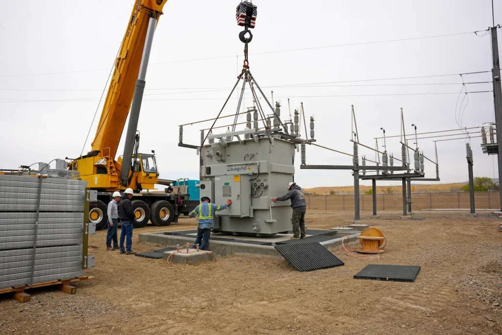 Workers guide a large electrical transformer being positioned by a crane at an outdoor power substation.