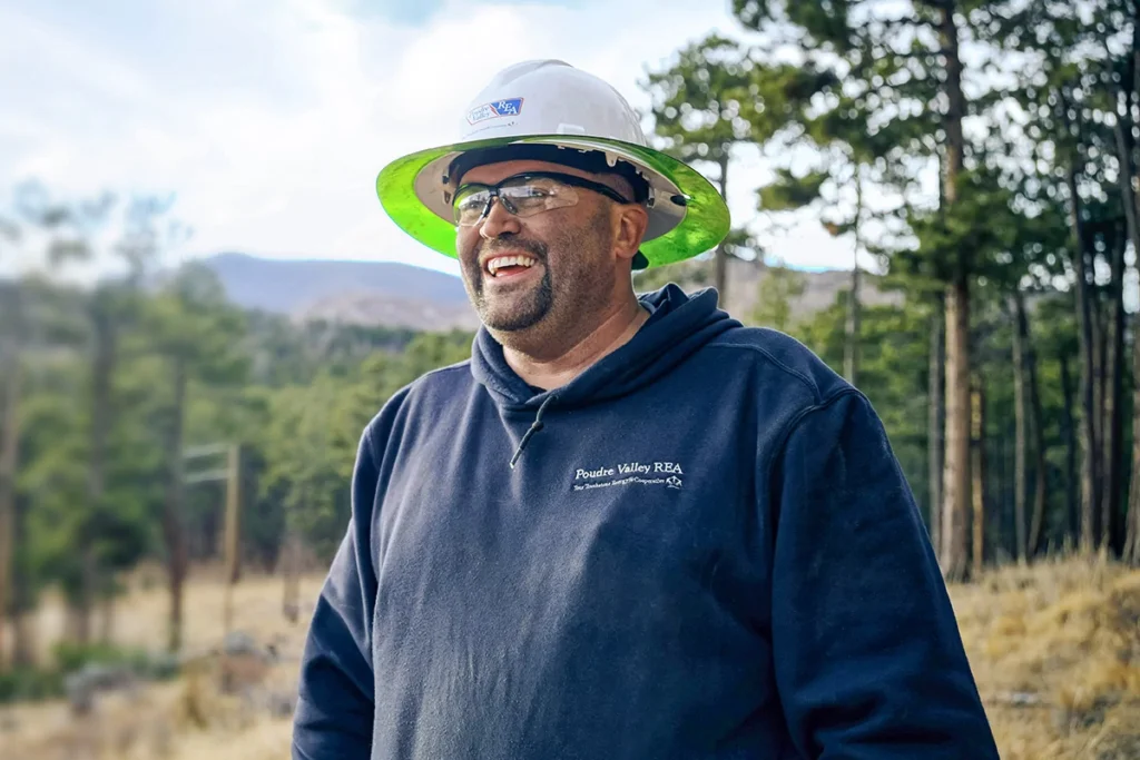A man wearing a hard hat and safety glasses smiles outdoors in a wooded area, dressed in a dark hoodie with "Poudre Valley REA" printed on it.