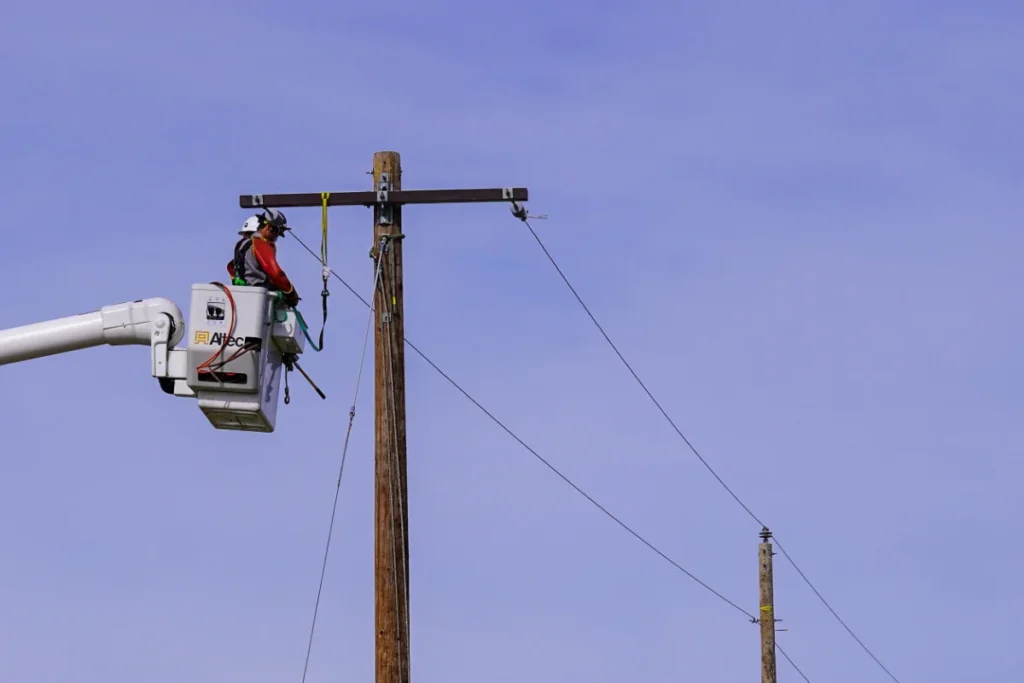 A utility worker in a bucket truck repairs or installs power lines on a wooden utility pole against a clear blue sky.