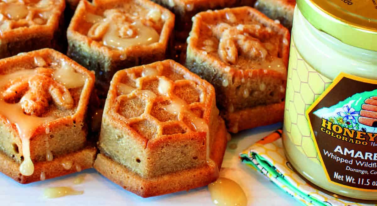 Hexagonal honey cakes with drizzled glaze and nut toppings are displayed beside a jar of whipped honey.