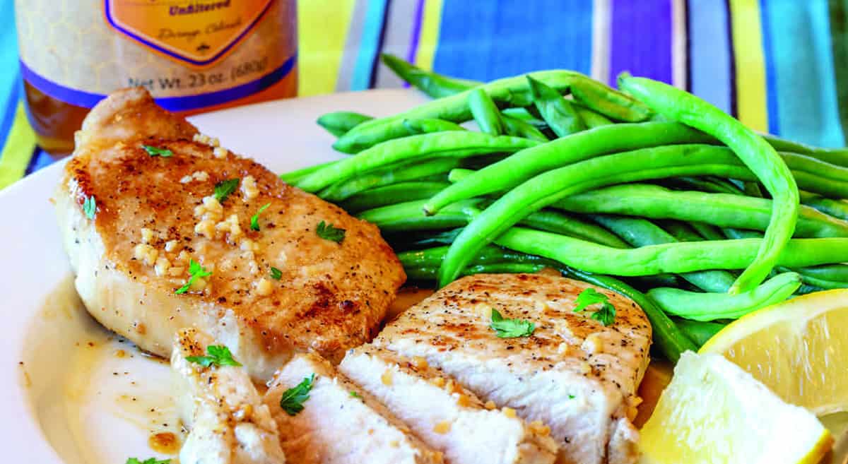 Grilled chicken breast with herbs, green beans, a lemon wedge, and a sauce bottle in the background on a white plate.