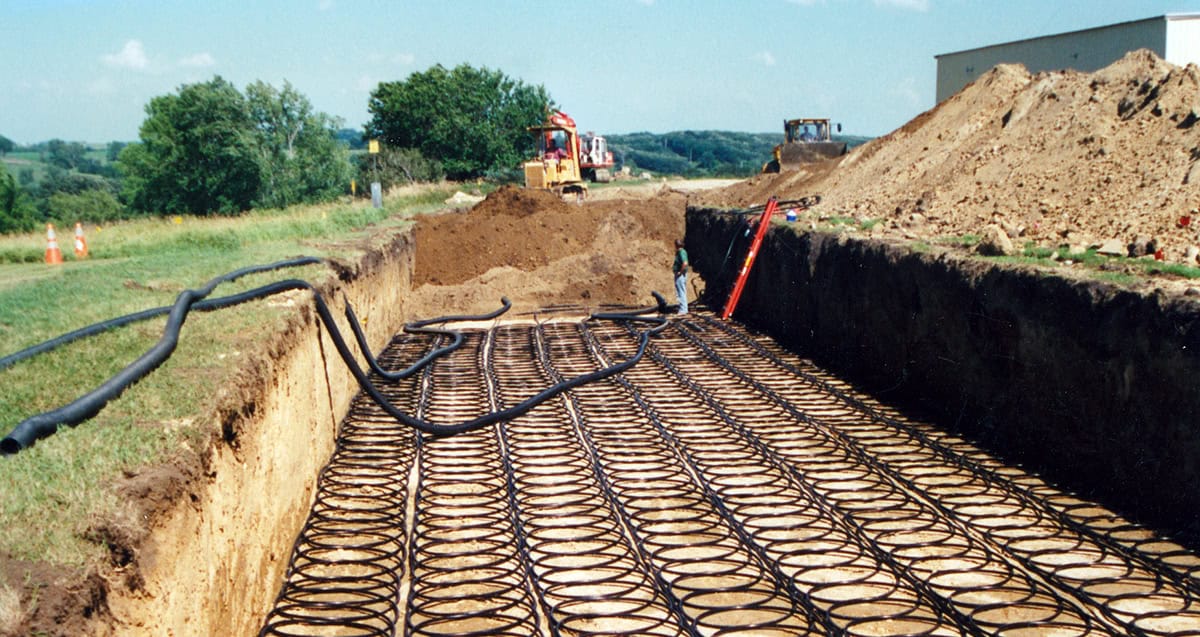 Construction site with long trench containing metal coil structures, surrounded by dirt piles. Machinery and trees visible in the background.