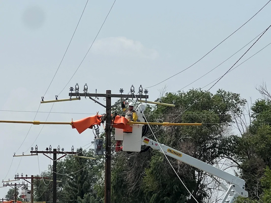 A utility worker in a bucket truck repairs power lines with safety equipment and orange protective covers on a clear day. Trees and multiple power poles are visible in the background.