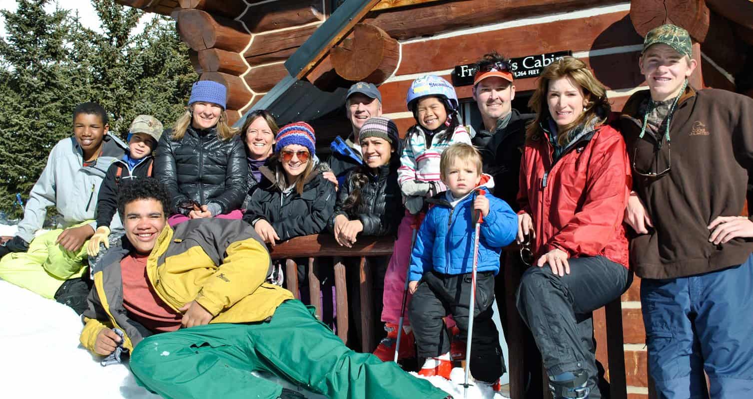 Group of people in winter clothing pose in front of a log cabin named "Fran's Cabin," surrounded by snow and trees.