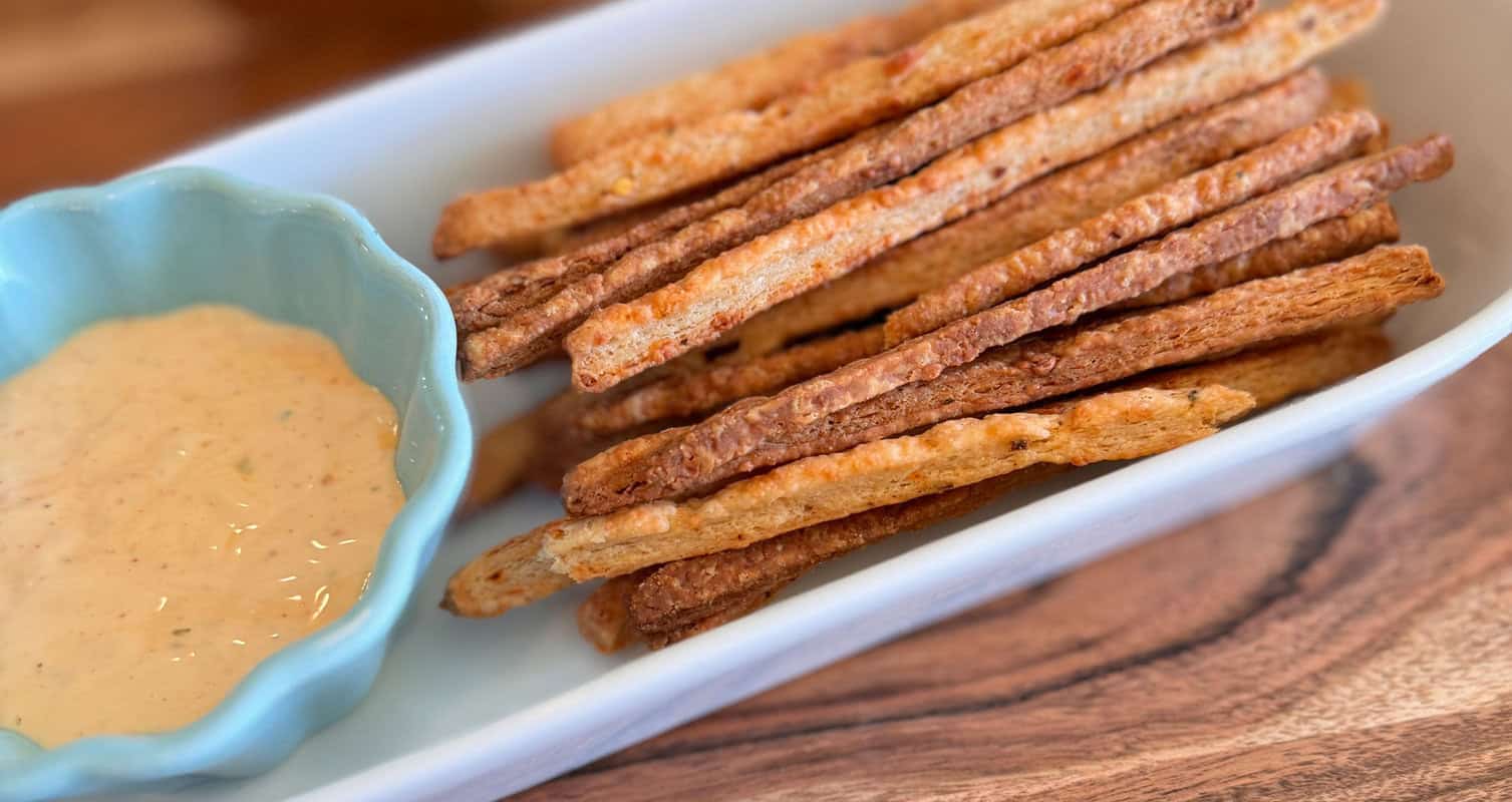 A rectangular white dish holds crispy breadsticks beside a small blue bowl filled with a creamy dipping sauce on a wooden surface.